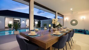 Un espace repas moderne avec une table en bois dressée pour six personnes, entourée de chaises, à proximité d’une piscine et d’un salon agrémenté de coussins turquoise.