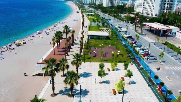 Un paseo marítimo frente a la playa con zona de estacionamiento, palmeras y vista al océano.