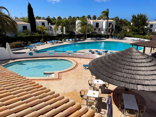 A resort pool area with lounge chairs and umbrellas, surrounded by palm trees and buildings.