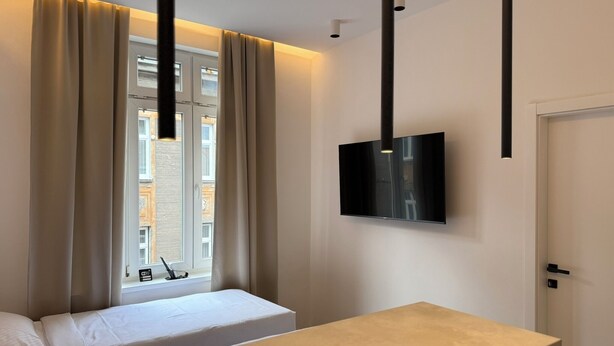 Ein modernes Hotelzimmer mit Bett, kleinem Tisch, Wandfernseher und einem Fenster mit Vorhängen.