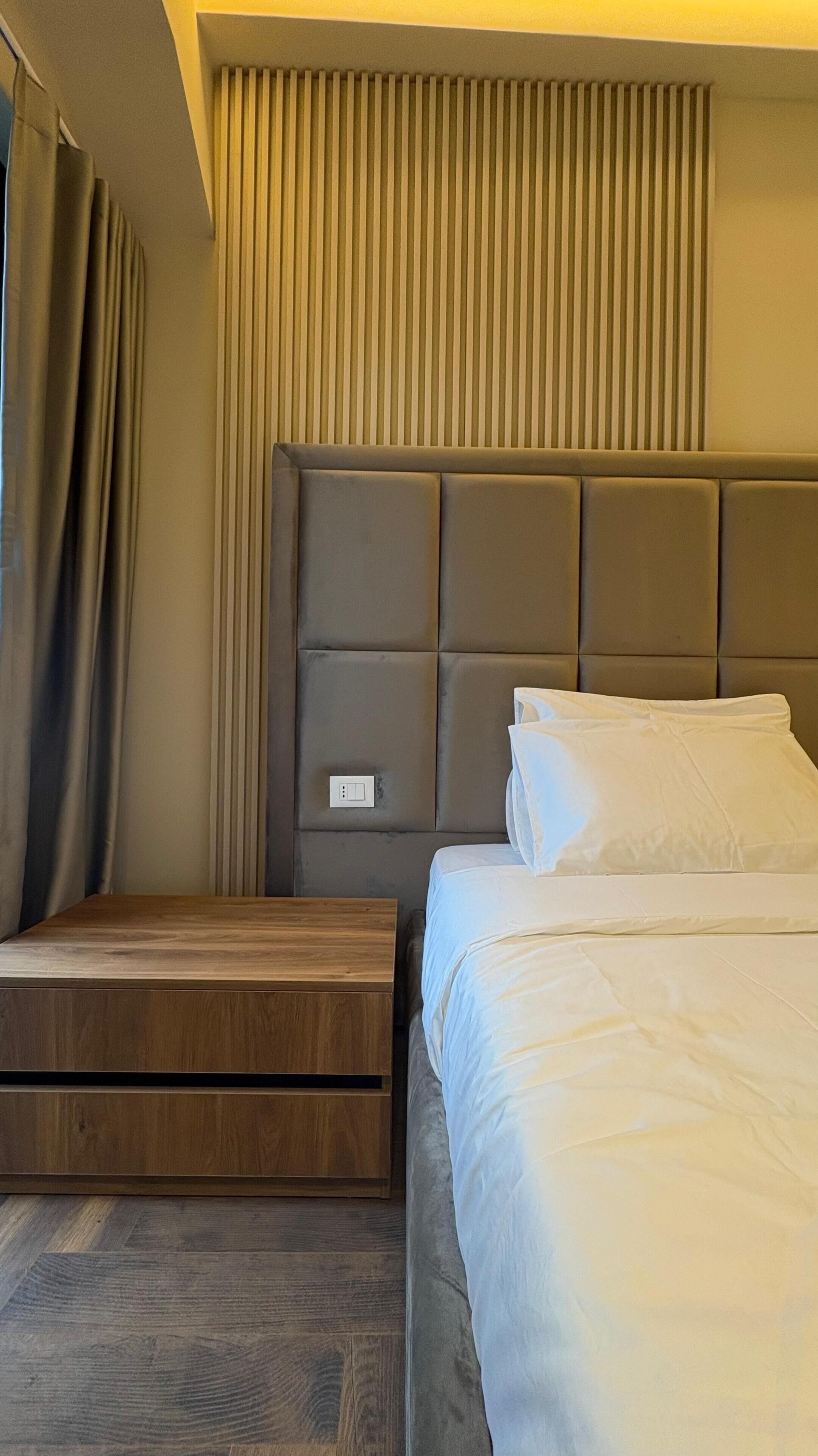 A hotel room with a bed, a wooden nightstand, a headboard with vertical slats, and a window with curtains.