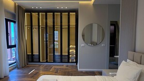 Egy modern szállodai szoba, amelyben egy nagy ágy, egy gardróbszerű beépített szekrény, egy kerek tükör található, és kilátás nyílik a külső környezetre.