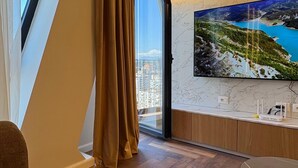 Egy modern nappali, melyben egy nagy márványfalra szerelt televízió, parketta és egy nagy ablakon át nyíló városi panoráma található.