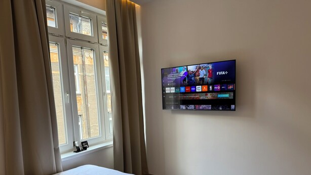 A hotel room with two beds, a TV mounted on the wall displaying various channels, and a window with curtains.