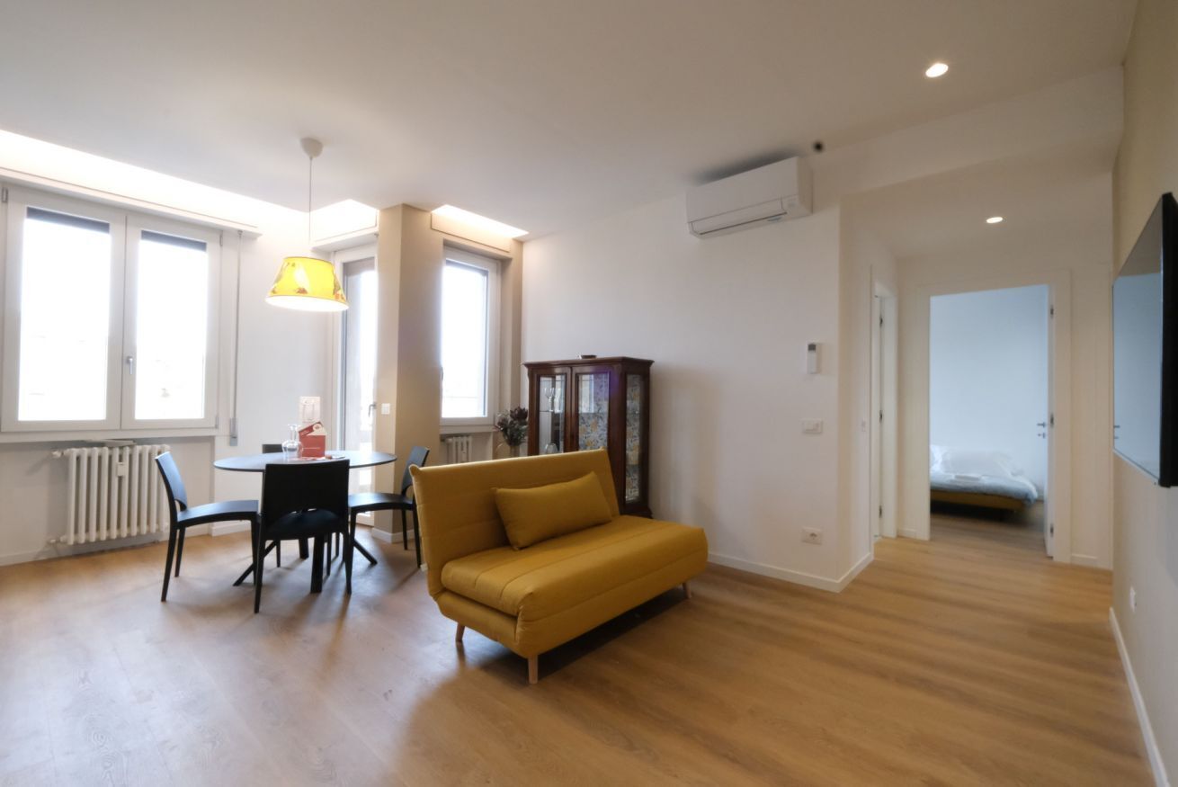 A modern living room with a sofa, dining table, and chairs. There is a window with a lamp, a radiator, and a cabinet.