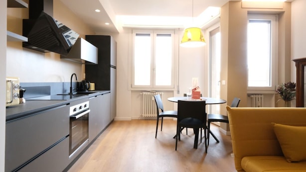 Une cuisine moderne équipée d’une cuisinière, d’un four et de meubles de rangement noirs. On y trouve un coin repas avec une table et des chaises, ainsi qu’un canapé jaune.