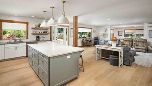 Cozinha espaçosa com uma ilha grande, armários de madeira e iluminação pendente. Há uma área de jantar com mesa e cadeiras, e uma sala de estar com sofá e mesa de centro.