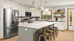 Cozinha moderna com eletrodomésticos de aço inoxidável, uma ilha grande com banquetas, iluminação pendente e uma janela com vista para áreas verdes.