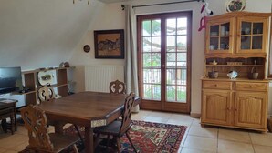 Una zona de menjador amb taula i cadires de fusta, televisió, un armari amb portes de vidre i una finestra amb vista a vegetació.