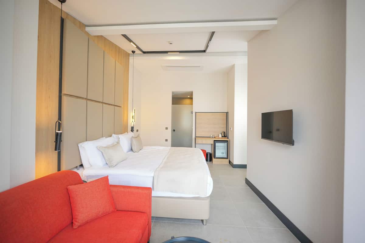 A modern hotel room with a large bed, a red sofa, a TV mounted on the wall, and a kitchenette area.