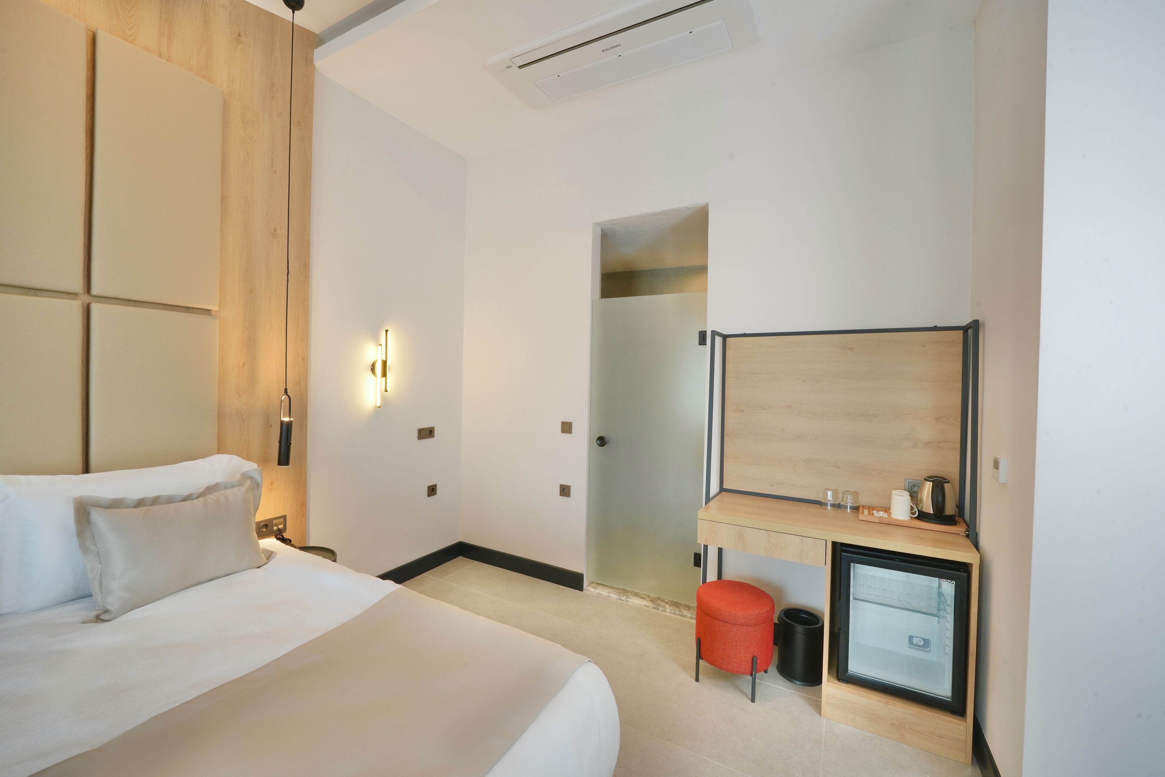 A modern hotel room with a large bed, a desk with a computer, a mirror, and a small stool.