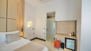 A modern hotel room with a large bed, a desk with a computer, a mirror, and a small stool.