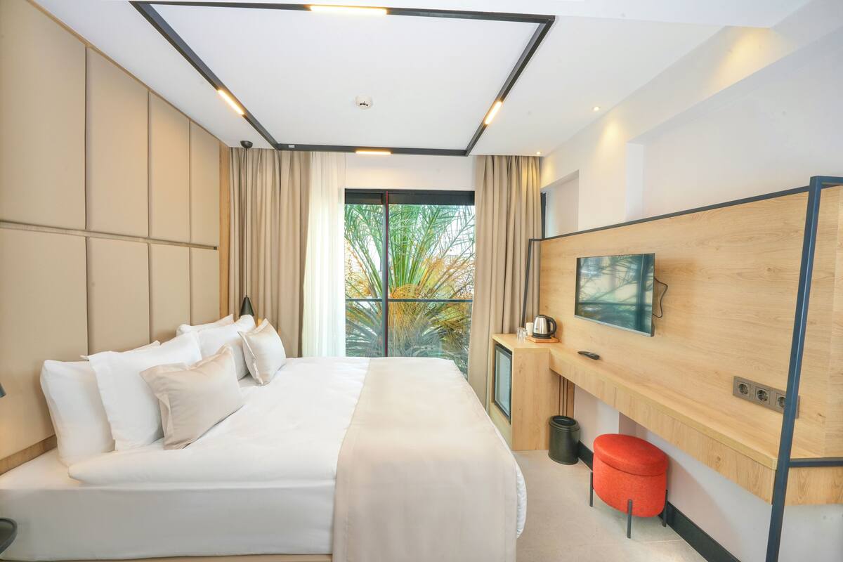 A modern hotel room with a large bed, a desk with a computer, a TV mounted on the wall, and a window with a view of trees.
