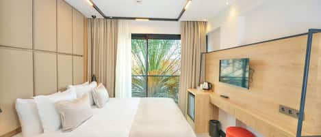 A modern hotel room with a large bed, a desk with a computer, a TV mounted on the wall, and a window with a view of trees.