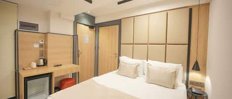 A hotel room with a bed, a desk with a coffee maker, a mini fridge, a wardrobe, and a bedside table with a lamp.