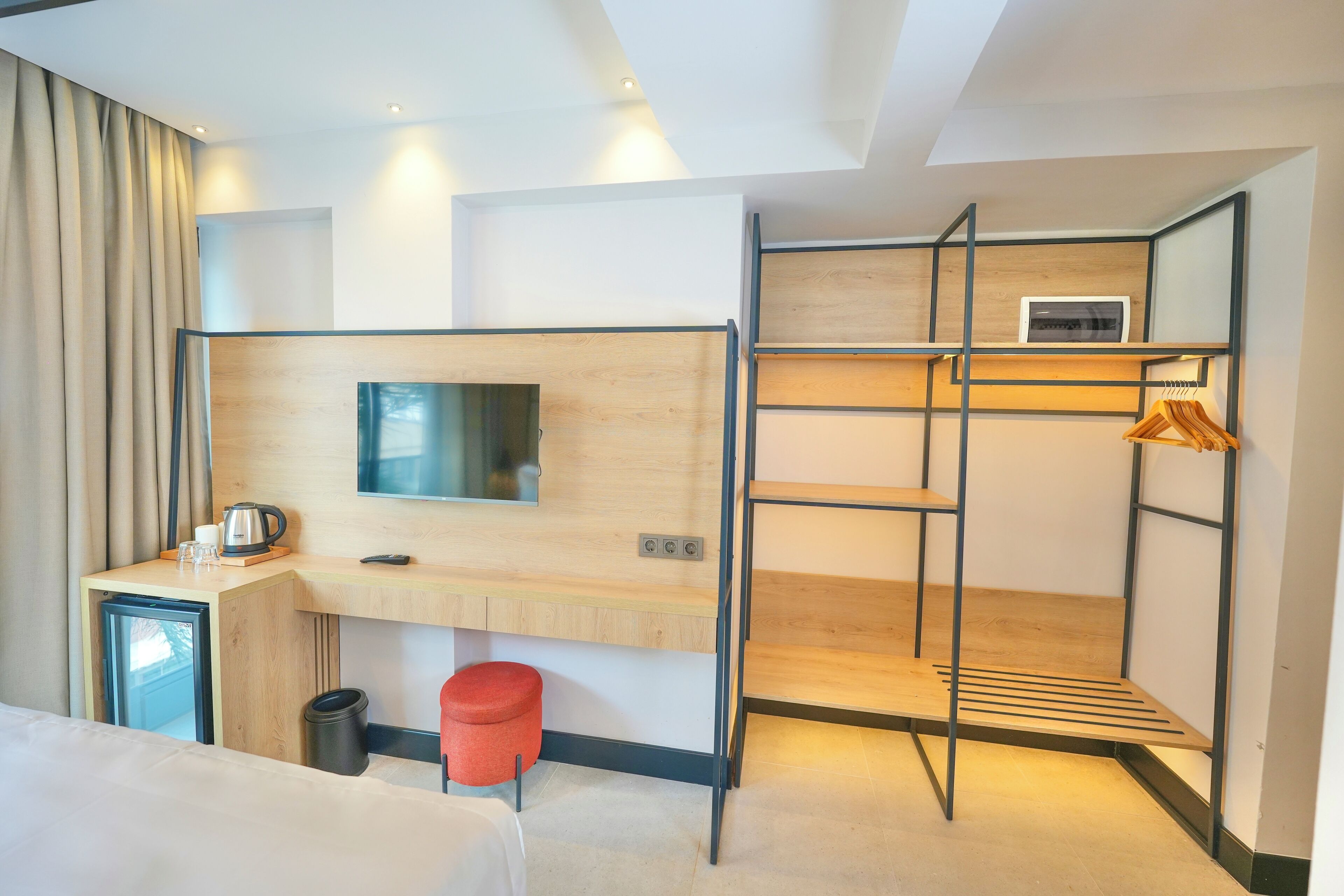 A compact hotel room with a bed, a small desk with a kettle, a TV, a mini-fridge, a red trash bin, and a built-in wardrobe with hanging space.