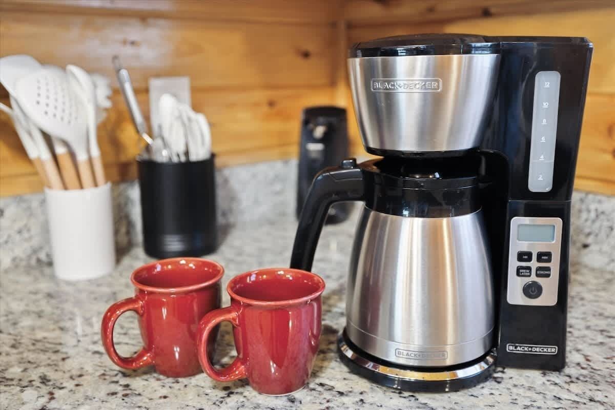 A coffee maker with a digital control panel, two red mugs, and a utensil holder on a kitchen counter.