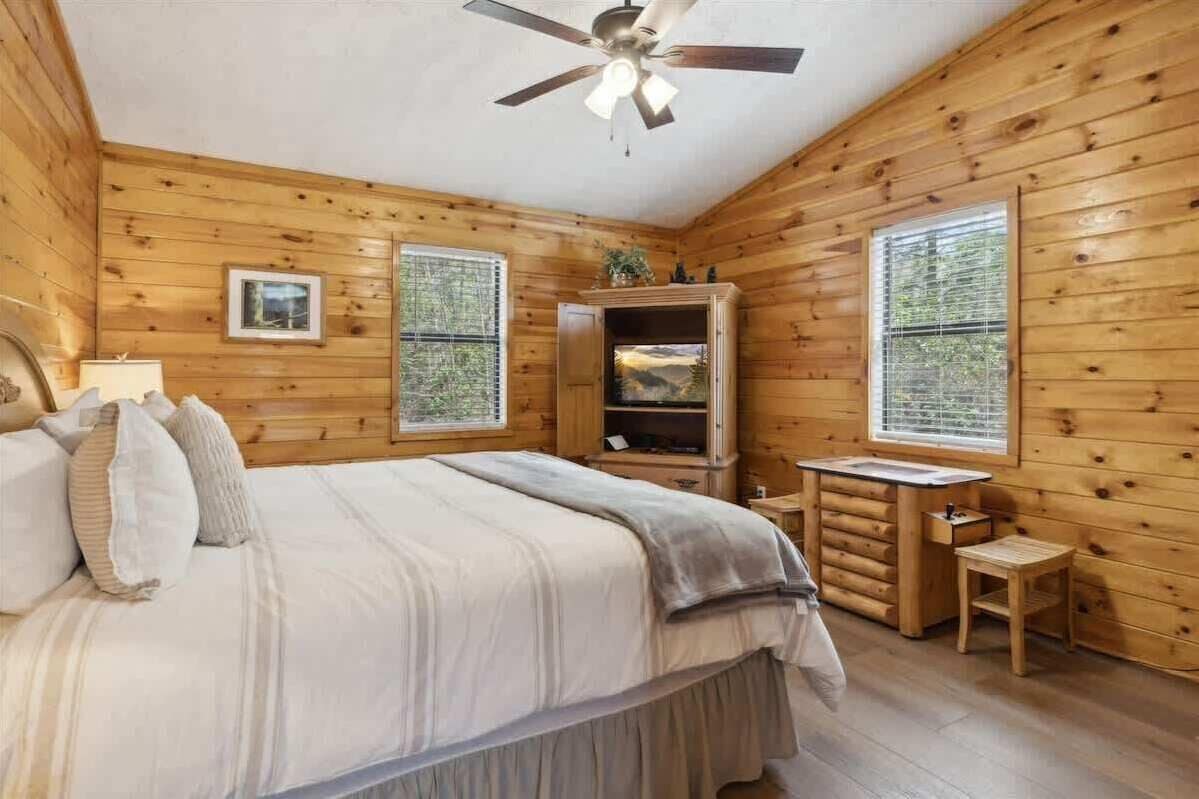 A wooden cabin bedroom with a bed, a ceiling fan, a small table, and a window with blinds.