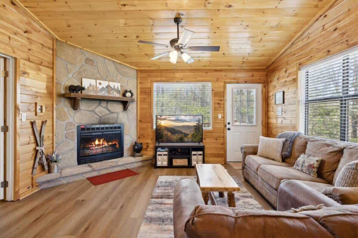 A cozy cabin interior with a stone fireplace, a television, wooden walls, and a wooden ceiling.
