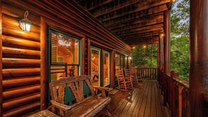 A wooden cabin with a porch, wooden benches, and a view of the forest.