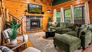 A cozy log cabin interior with a stone fireplace, green upholstered furniture, and a television mounted on the wall.