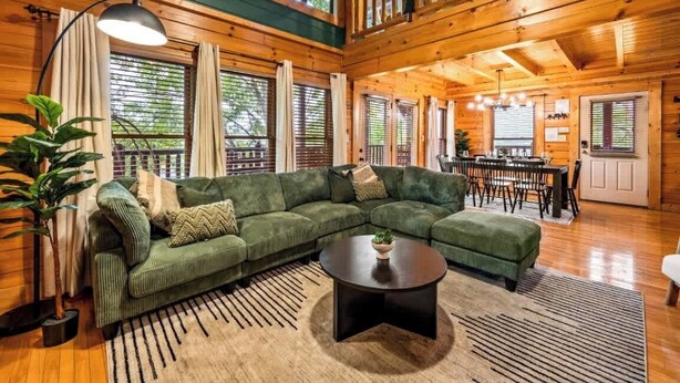 A cozy log cabin interior with a green sectional sofa, a round coffee table, and a dining area with a wooden table and chairs.