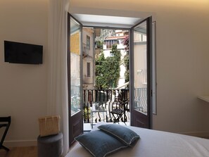 A bedroom with a bed, pillows, a chair, and a small table. There is a balcony with a railing and a view of buildings and trees.