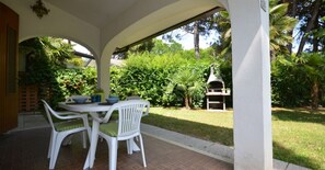 Une terrasse couverte avec une table et des chaises, entourée d’une végétation luxuriante et agrémentée d’un barbecue.