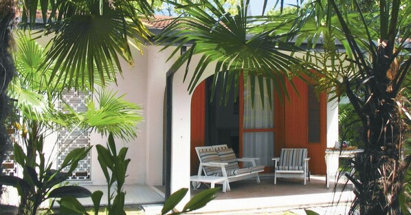 Un espace extérieur aménagé avec une zone de sièges couverte, comprenant deux chaises blanches et une table, le tout entouré d’une végétation tropicale luxuriante.