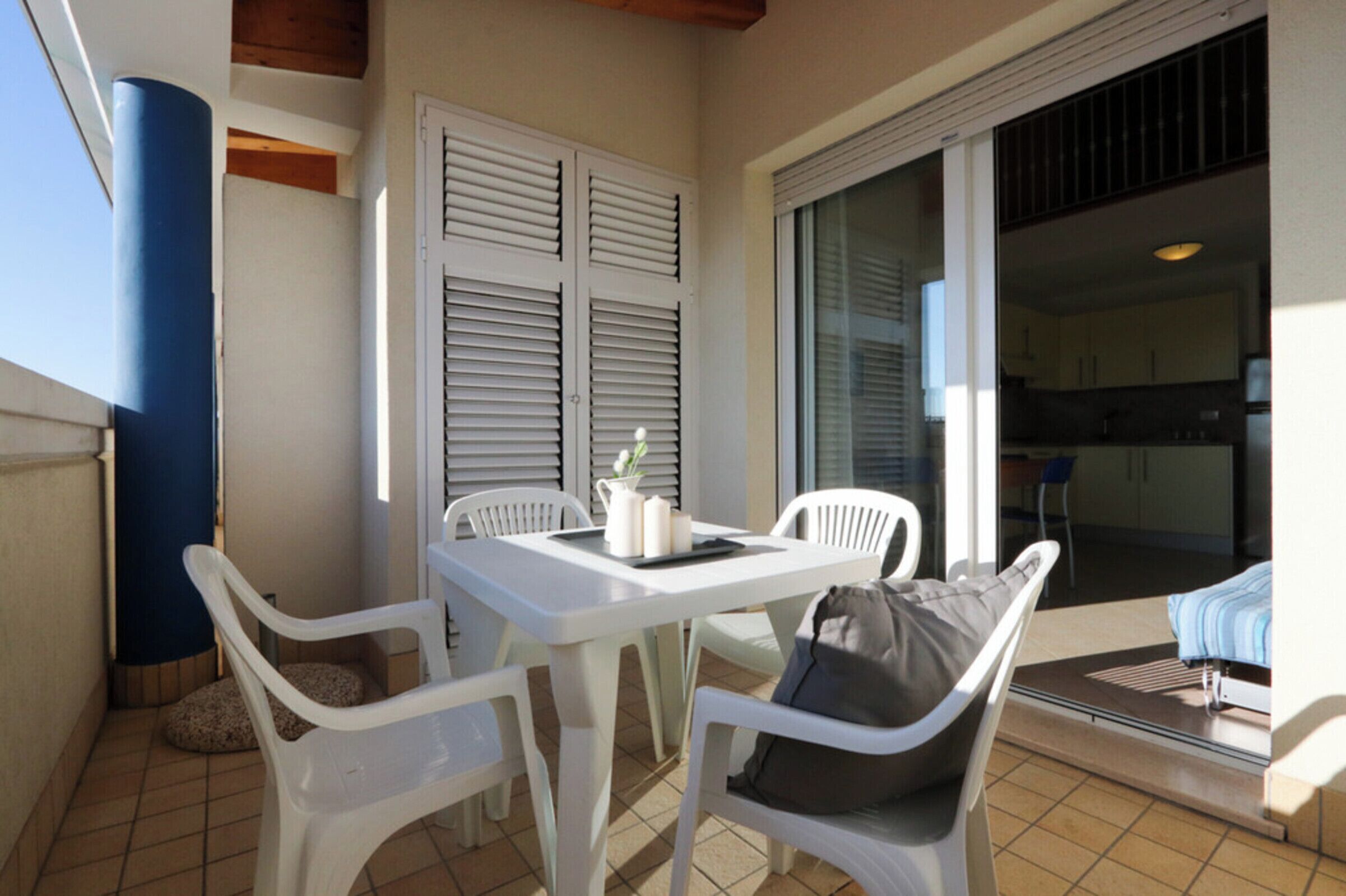A balcony with a white table and chairs, a blue pillar, and a sliding glass door leading to a kitchen area.