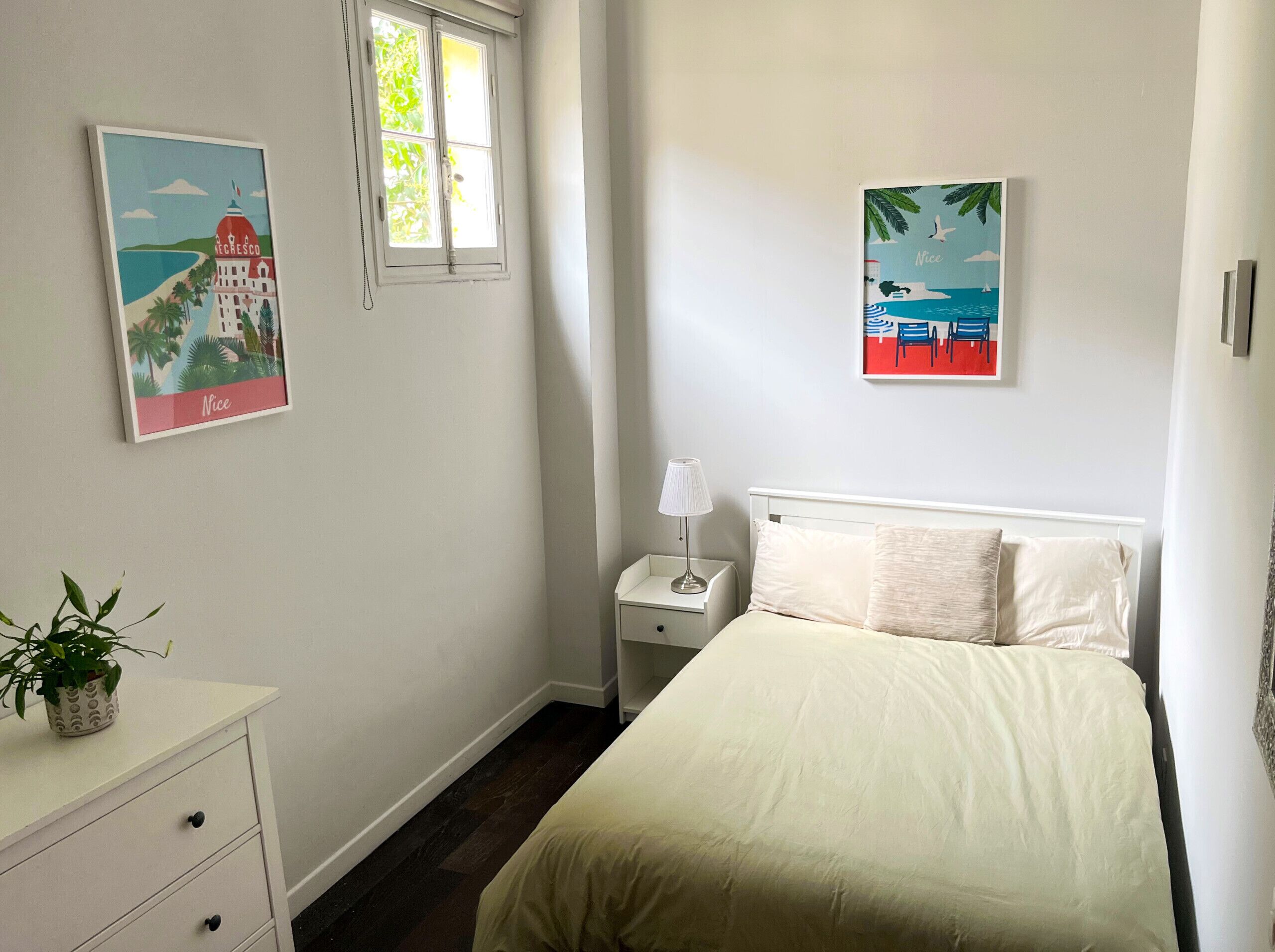 A bedroom with a bed, a nightstand, a lamp, a potted plant, and two wall art pieces.