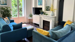 A living room with a blue sofa, a small white table, a TV, a fireplace, and a large window with plants outside. - Berlioz Blue AP4458 by Riviera Holiday Homes (Niza)