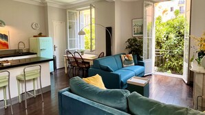 A modern living room with a blue sofa, a dining area with a table and chairs, a kitchen with a fridge, and a view of greenery through a glass door. - Berlioz Blue AP4458 by Riviera Holiday Homes (Niza)