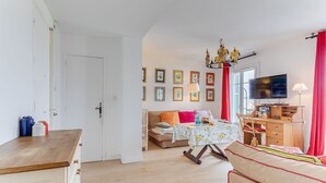 Un salon avec un canapé, une table à manger et un téléviseur. Les murs sont ornés de tableaux encadrés et un lustre est suspendu au plafond.
