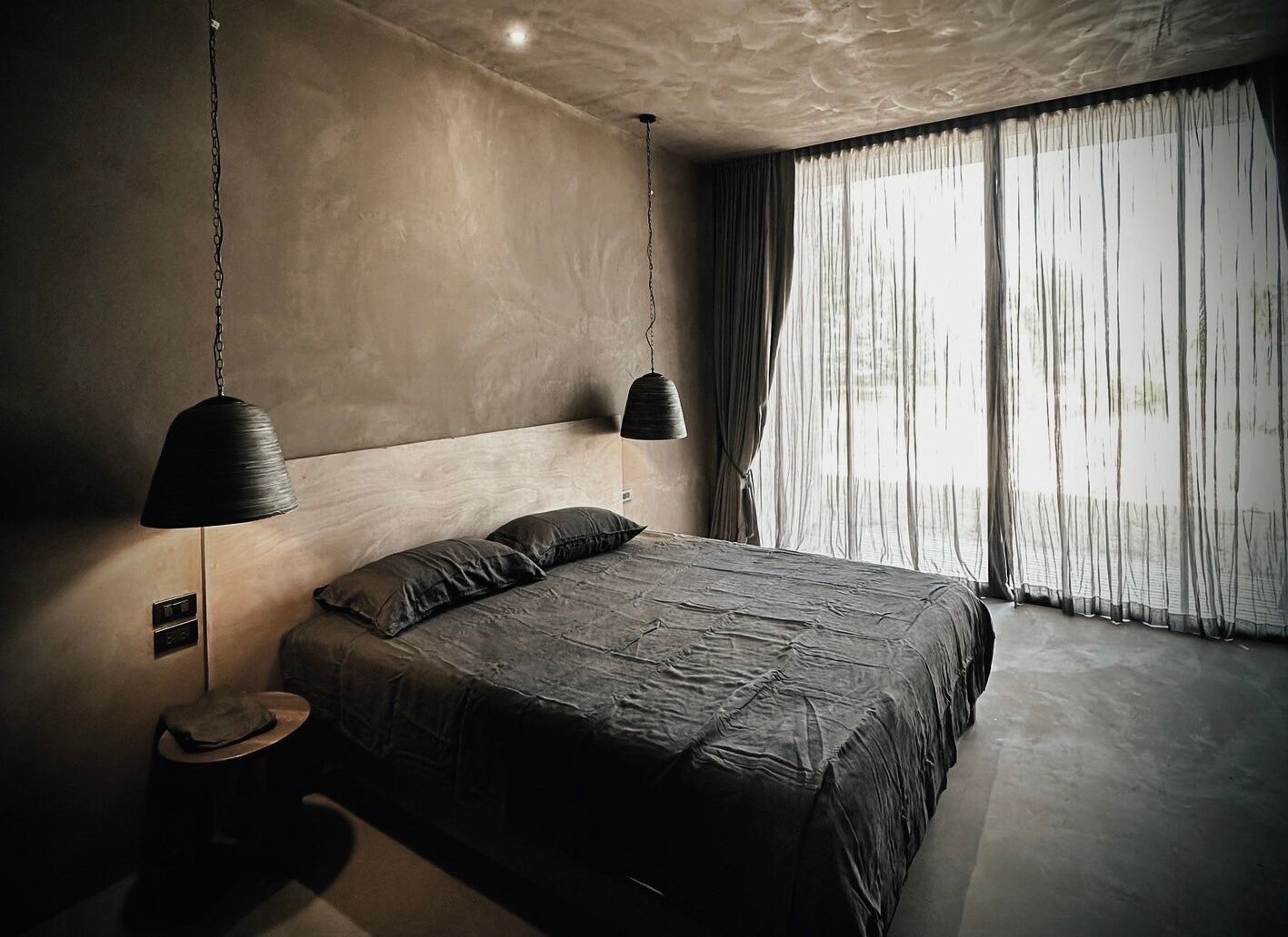 Une chambre moderne avec un grand lit, deux lampes suspendues et une vue sur l’extérieur à travers des rideaux légers.