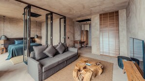 Ein modernes Wohnzimmer mit einem grauen Sofa, einem Couchtisch mit Holzscheiten und einem großen Fenster mit Holzjalousien.