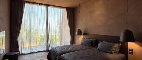A modern bedroom with a large bed, a desk, and a view of the outdoors through sheer curtains.