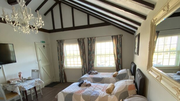 A room with a high ceiling, exposed beams, a chandelier, a bed with a quilt, a sofa with cushions, a television, and a window with curtains.