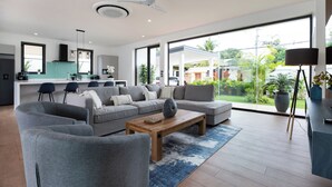 A modern living room with a sofa, coffee table, and chairs. There is a kitchen area with a dining table and chairs. Large windows provide a view of the outdoors.