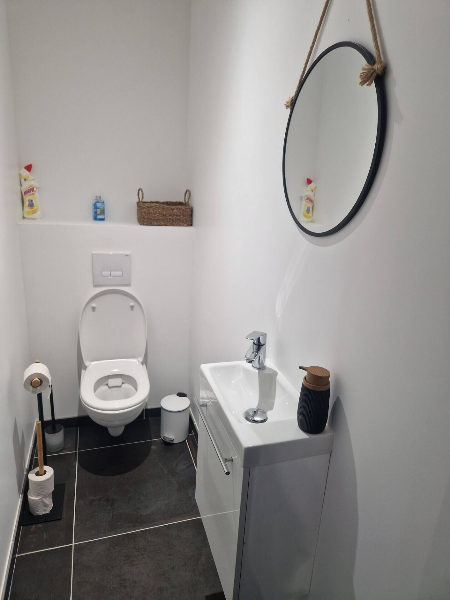 Een compacte, moderne badkamer met toilet, wastafel en spiegel. Er is een plank met toiletartikelen en een klein mandje.