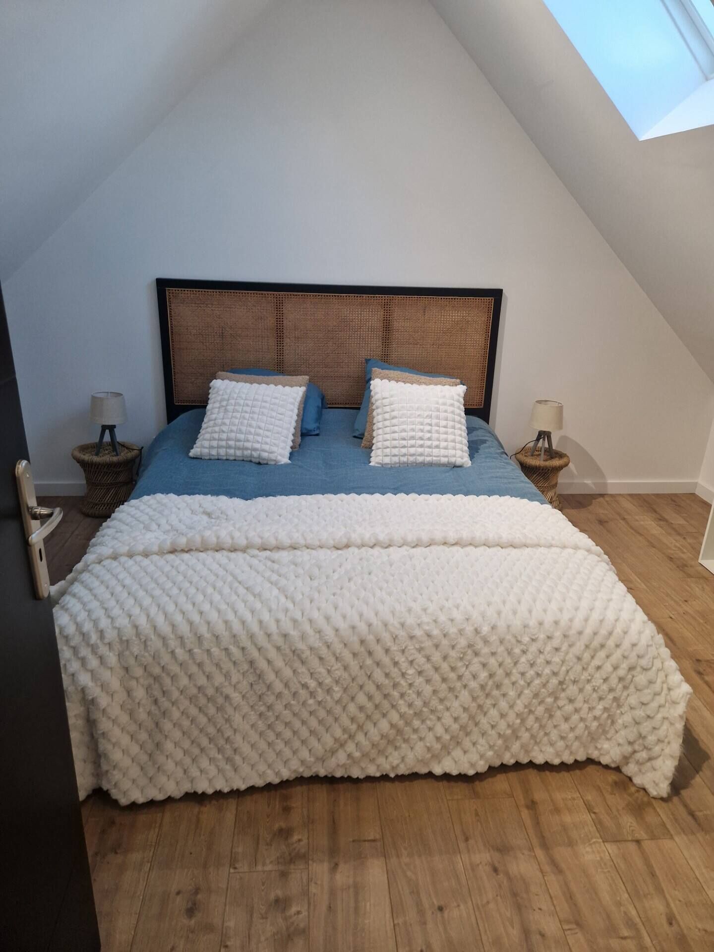 A bedroom with a sloped ceiling, a bed with a blue bedspread and white pillows, a white textured blanket, and two bedside lamps on wicker stands.