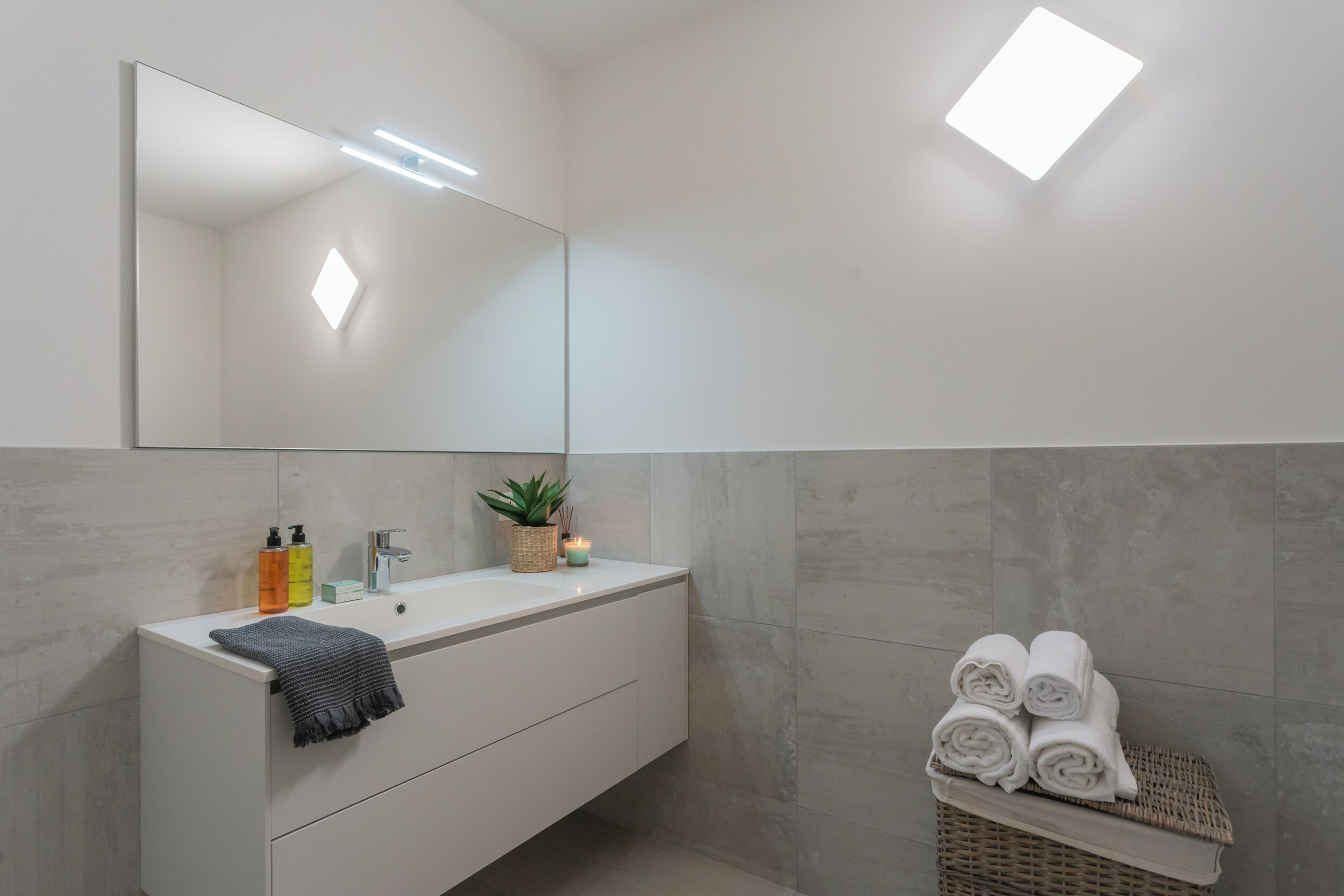 Un baño moderno con tocador blanco, un espejo grande y una lámpara empotrada. Hay artículos de aseo en el tocador y una cesta con toallas enrolladas.