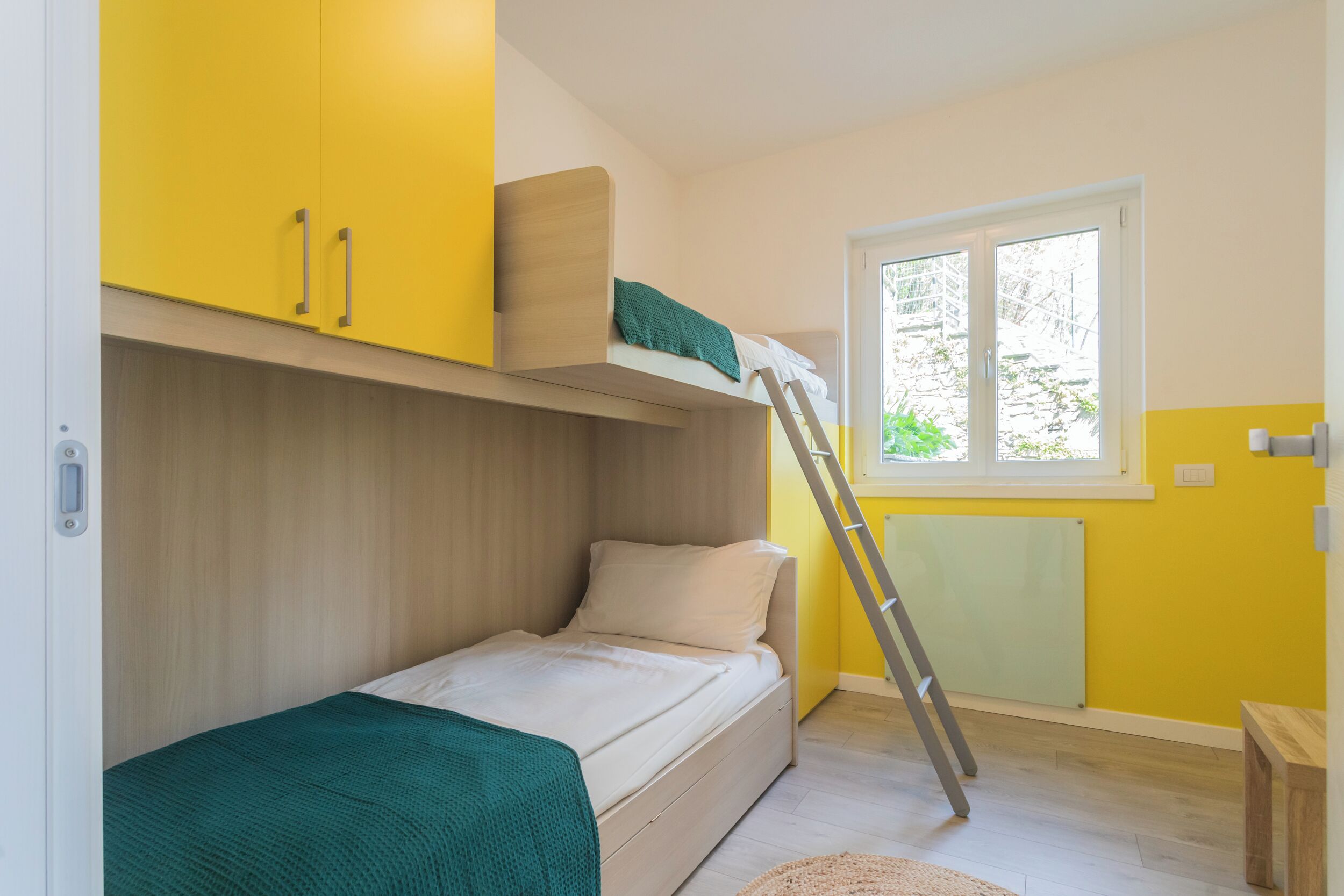 A small, compact room with a bunk bed, a window with a view of greenery, and a radiator.