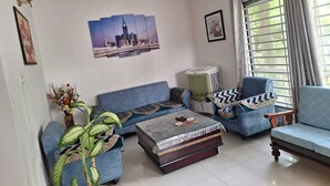 Ein Wohnzimmer mit einer Couch, einem Sessel und einem Couchtisch. Es gibt einen Deckenventilator, ein Fenster mit Vorhängen und eine Wand-Klimaanlage.