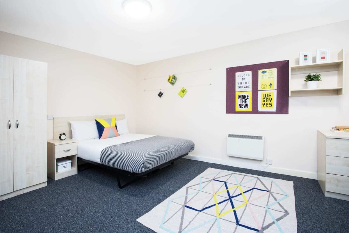 A modern bedroom with a bed, bedside table, and a geometric rug. There are wall decorations and a small shelf with items.