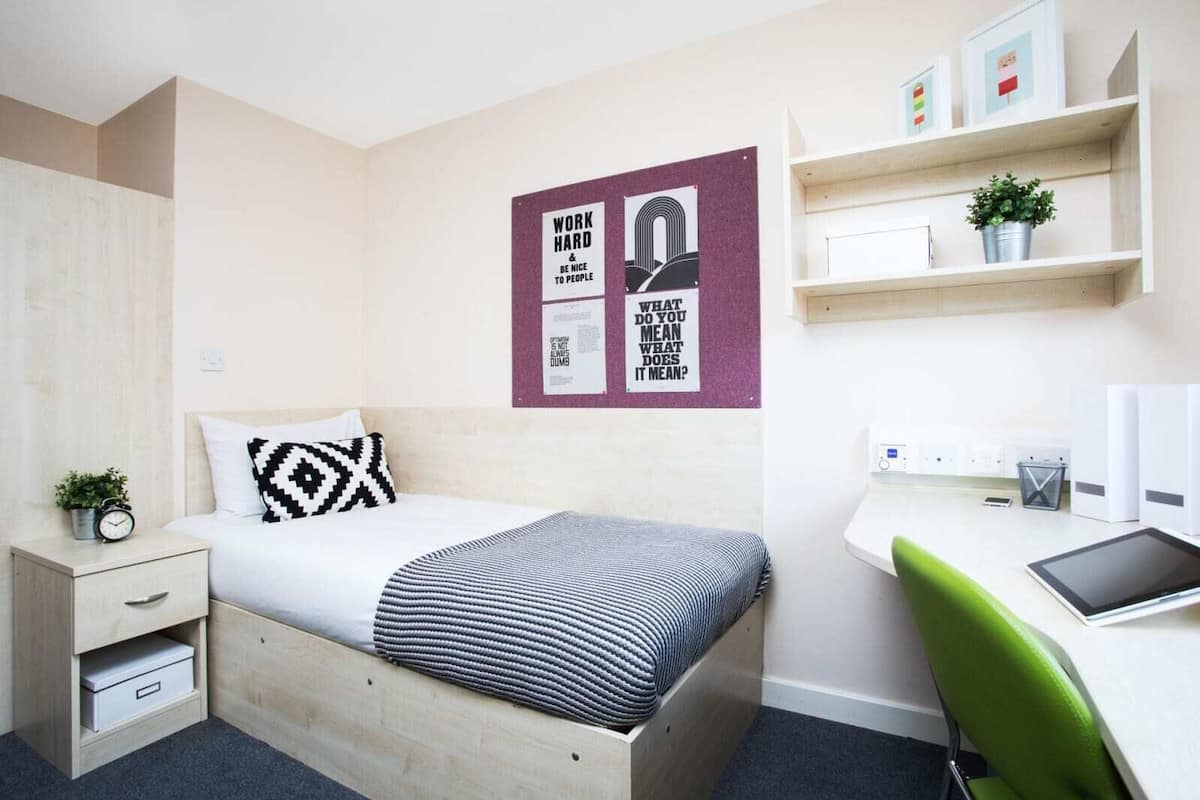 A modern bedroom with a bed, bedside table, and a desk with a chair. There are wall art pieces and a small plant on the desk.