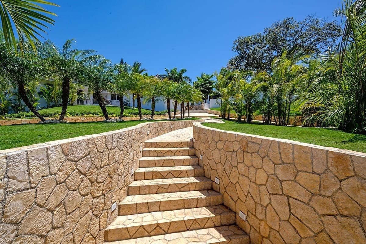 A stone staircase leading up to a building, surrounded by a well-maintained garden with palm trees and a clear blue sky.