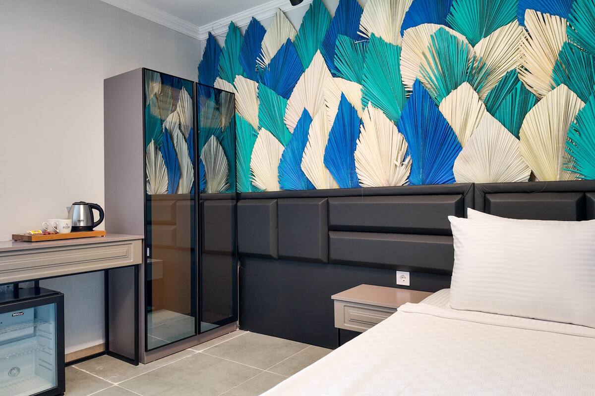 Habitación de hotel con cama, escritorio con cafetera, minibar y un decorado de pared con motivos de hojas.