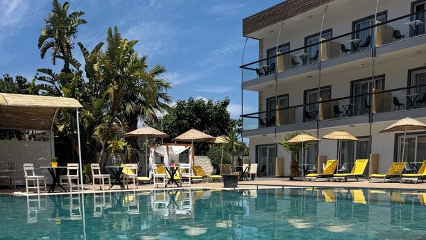 A clear pool with lounge chairs and umbrellas, surrounded by palm trees and a multi-story building. - Mos Hotel Çeşme (Cesme)
