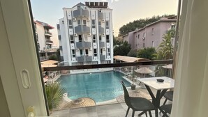 A view of a swimming pool with a tiled patio area, a table with chairs, and a multi-story building in the background. - Mos Hotel Çeşme (Cesme)
